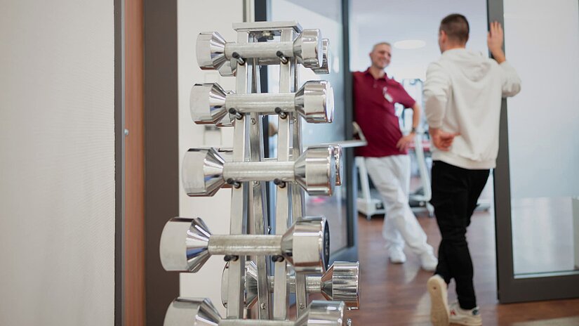 Im Vordergrund verschieden schwere Hanteln gestapelt auf einer Halterung und im Hintergrund ein Therapeut und ein Sportler bei der Erstberatung.